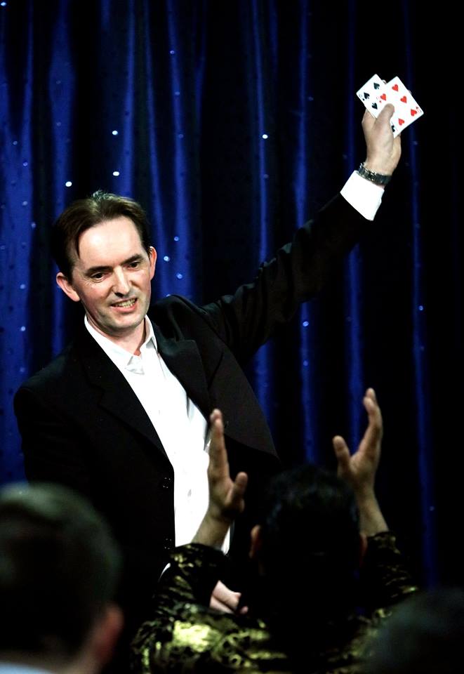 The Impossible Mr Goodwin on stage with an arm in the air holding two playing cards with the audience clapping